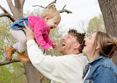 Joyful family portrait with a dad holding his giggling daughter up high while mum smiles beside them, in a beautiful park.