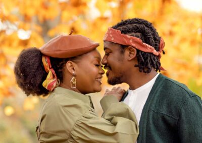 A couple with heads close together, almost touching noses, smiling at each other against a soft background of golden and orange autumn leaves. The woman wears a rust-colored beret and a green top, and the man has a red bandana on his head. The perfect autumnal portrait.