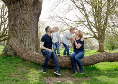 A playful family portrait captured outdoors, with parents and their two young children sitting on a fallen tree, exchanging kisses.